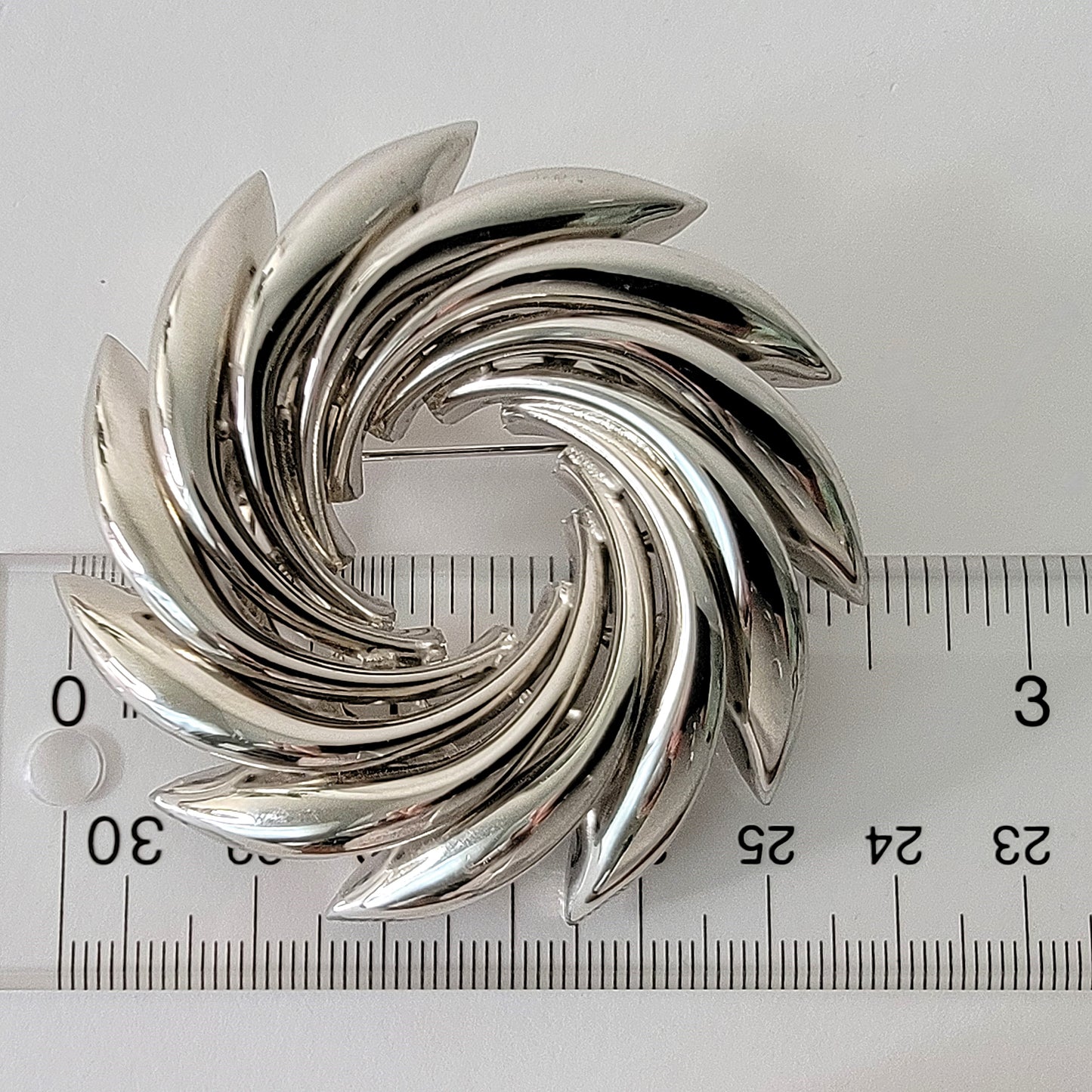 Silver tone vortex style modernist brooch next to a ruler.