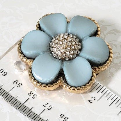 Light blue thermoset plastic style flower brooch, with glass rhinestones and a gold tone edge setting. Shown next to a ruler.