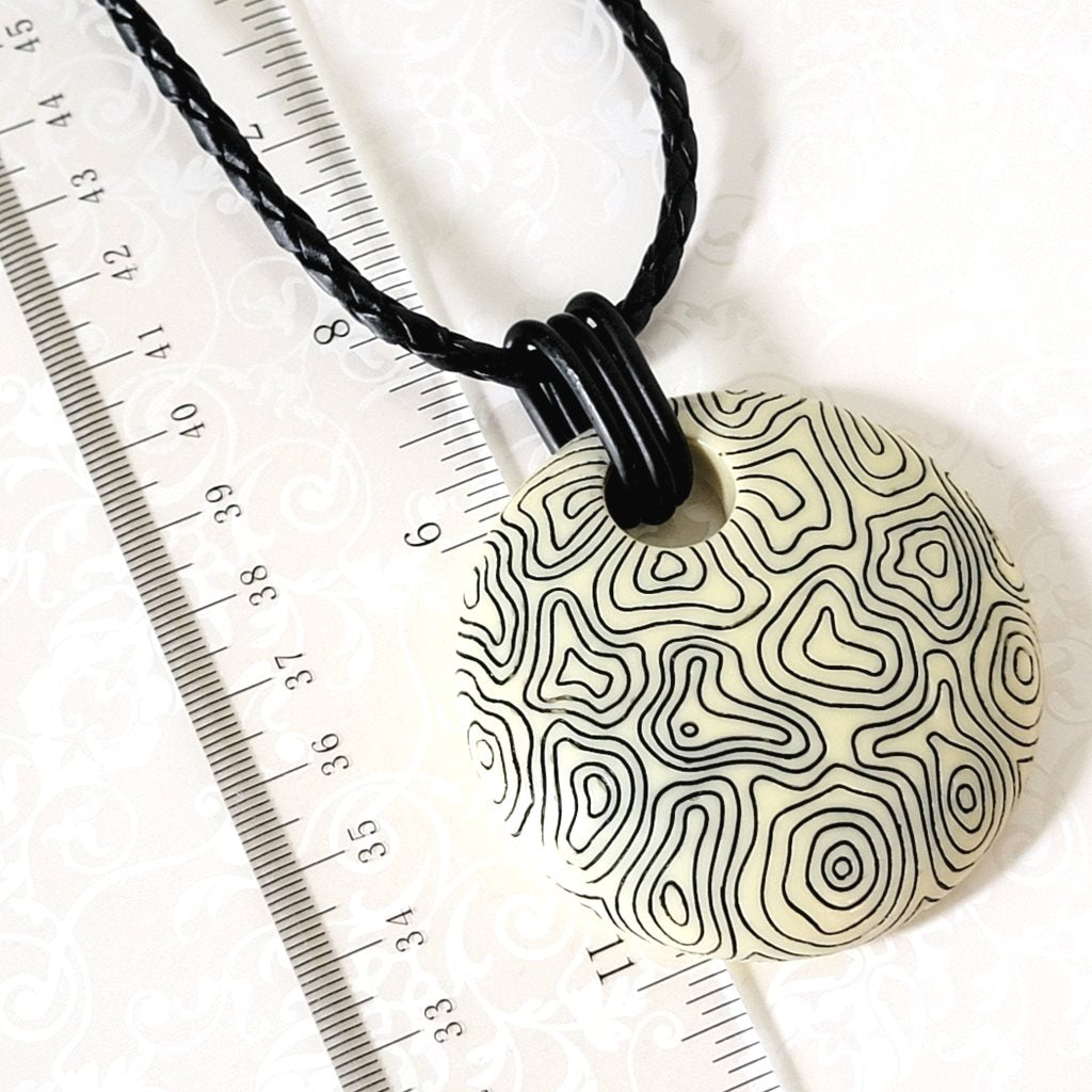 Closeup of large Chicos circle abstract pendant, with black swirling lines. Shown next to a ruler.