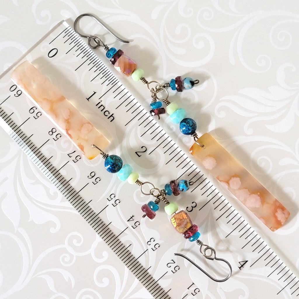 Long gemstone dangle earrings, in peach and blue colors, shown next to a ruler.