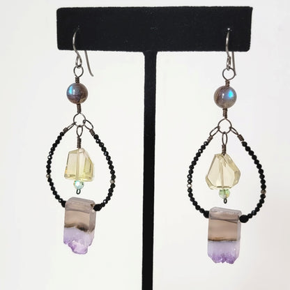Gemstone dangle hoop earrings, with amethyst slices, yellow quartz, and black spinel. Shown on a display stand.