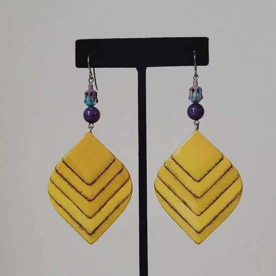 Video of a pair of large yellow wood dangle earrings, with purple and turquoise accents. Shown on a display stand.