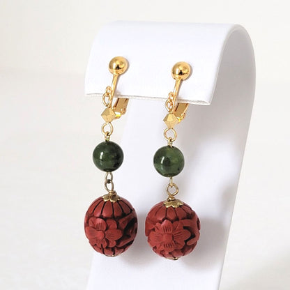 Floral cinnabar-look earrings with green stones, on gold tone clips, shown on a display stand.
