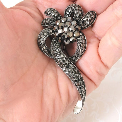 Silvery gray goth style rhinestone floral ribbon brooch, in antiqued silver tone setting, shown in hand, for size comparison.