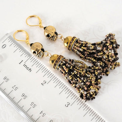 Long gold tone skull dangle bead tassel earrings, next to a ruler.